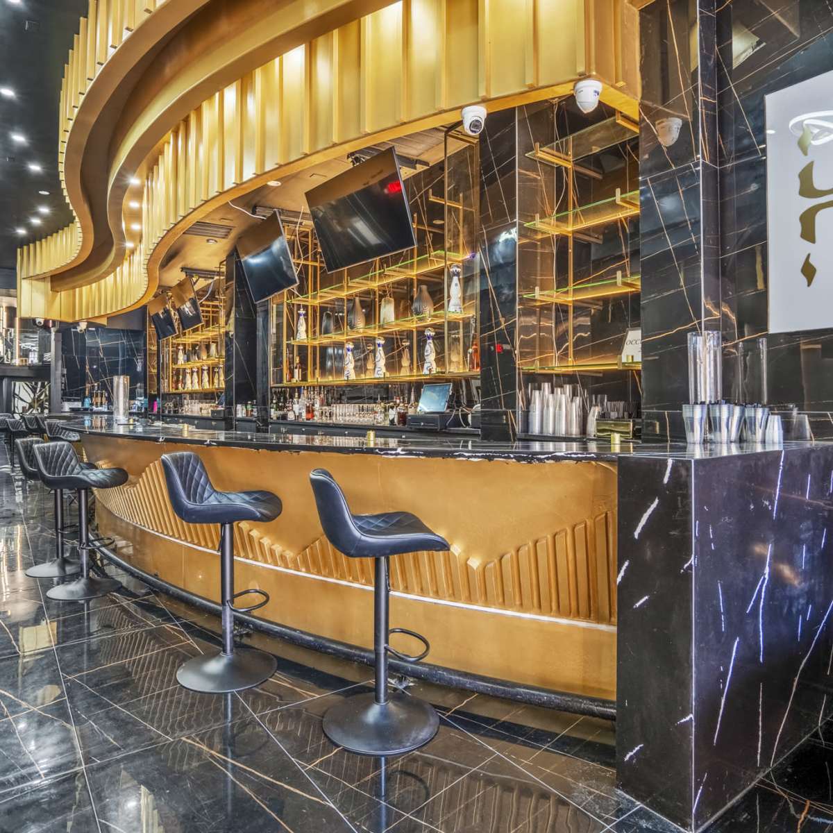 A luxurious bar with a golden counter and black marble flooring, featuring several bar stools and shelves stocked with various bottles.