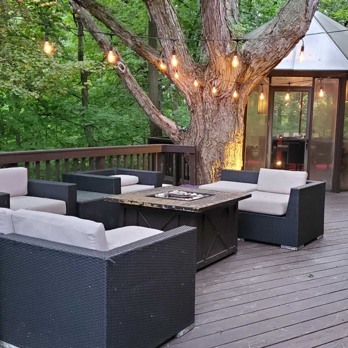 A wooden deck with four seating chairs arranged around a central table, decorated with string lights and surrounded by trees.