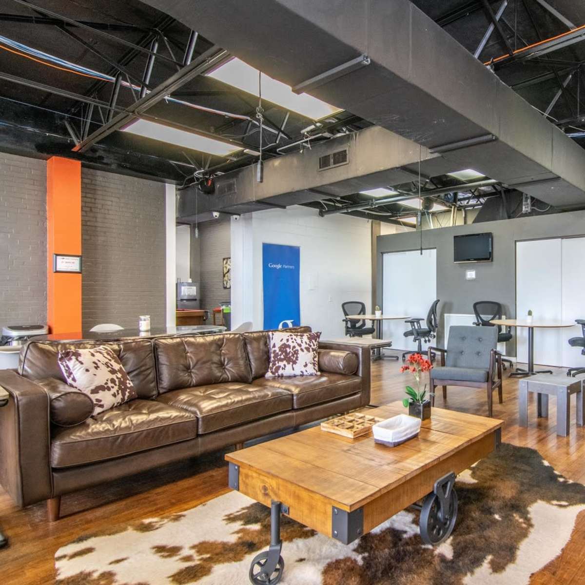 A modern office interior features a leather sofa, a wooden coffee table on a rug, and several workspaces with chairs and monitors in the background.