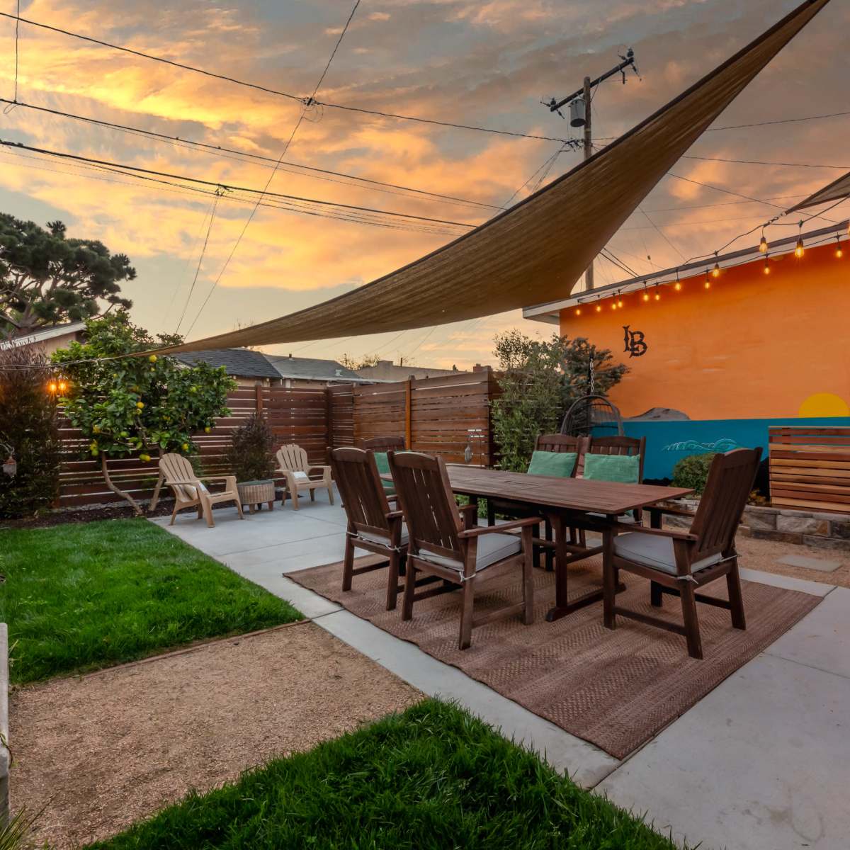 A landscaped outdoor area with a large dining table surrounded by chairs, string lights overhead, and a colorful mural on a nearby wall, set against a sunset sky.