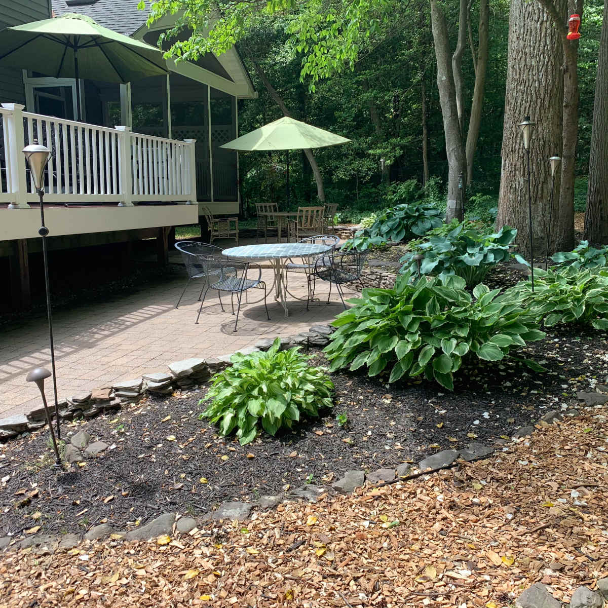 A circular stone patio with a table and chairs is surrounded by lush greenery and garden lights in a wooded area.