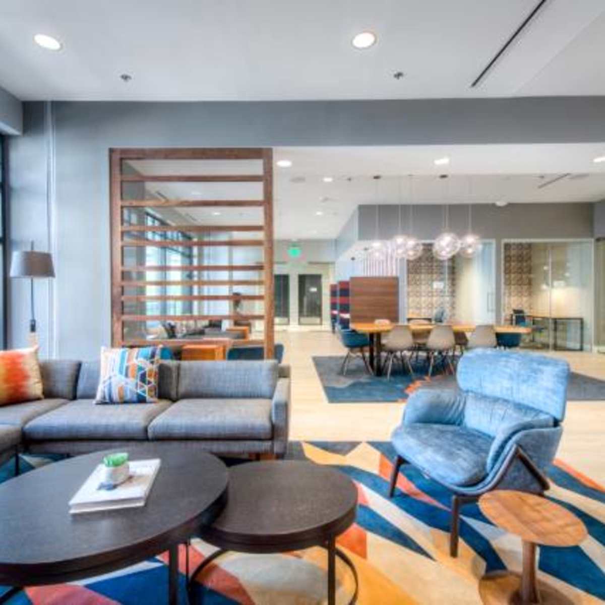 A modern lounge area with a sectional sofa, coffee tables, and a dining space featuring a large table and chairs, all arranged in a spacious, brightly lit room.