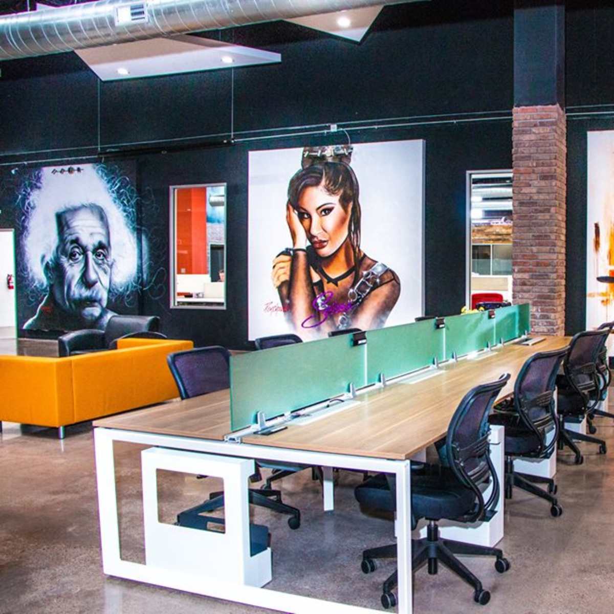 A modern workspace with multiple desks, black walls adorned with large mural artworks, and a view of vibrant orange and gray graffiti.
