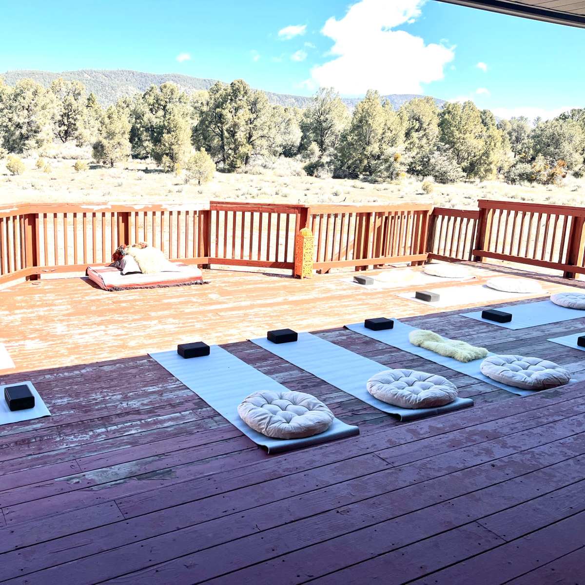 A spacious wooden deck with meditation cushions and mats arranged neatly, overlooking a landscape of trees and mountains.