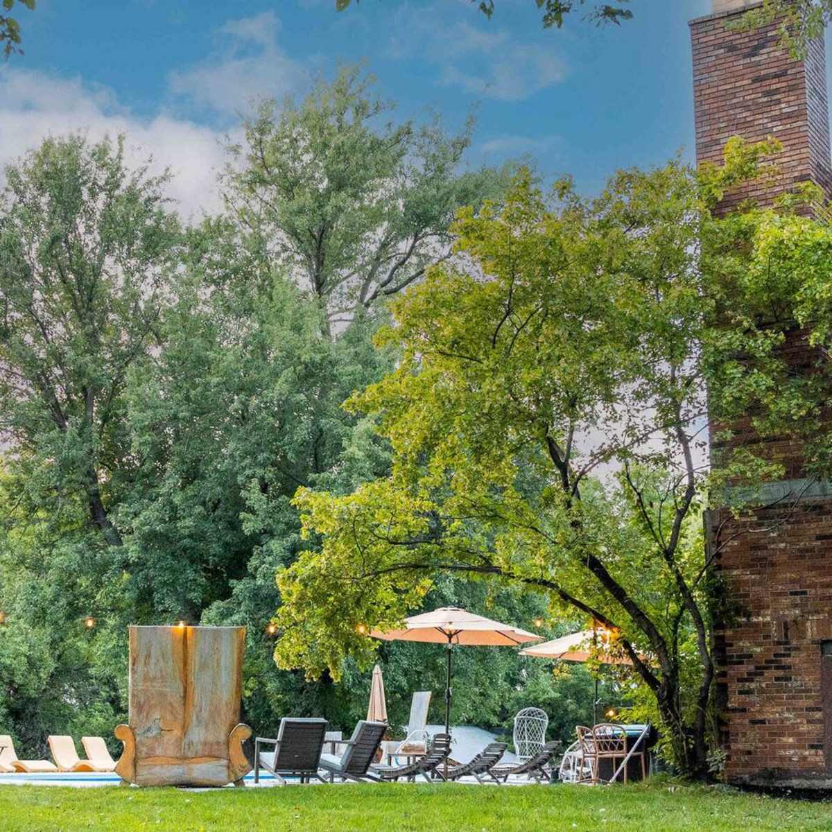 A lush outdoor area with lounge chairs, umbrellas, and a stone structure set against a backdrop of trees.