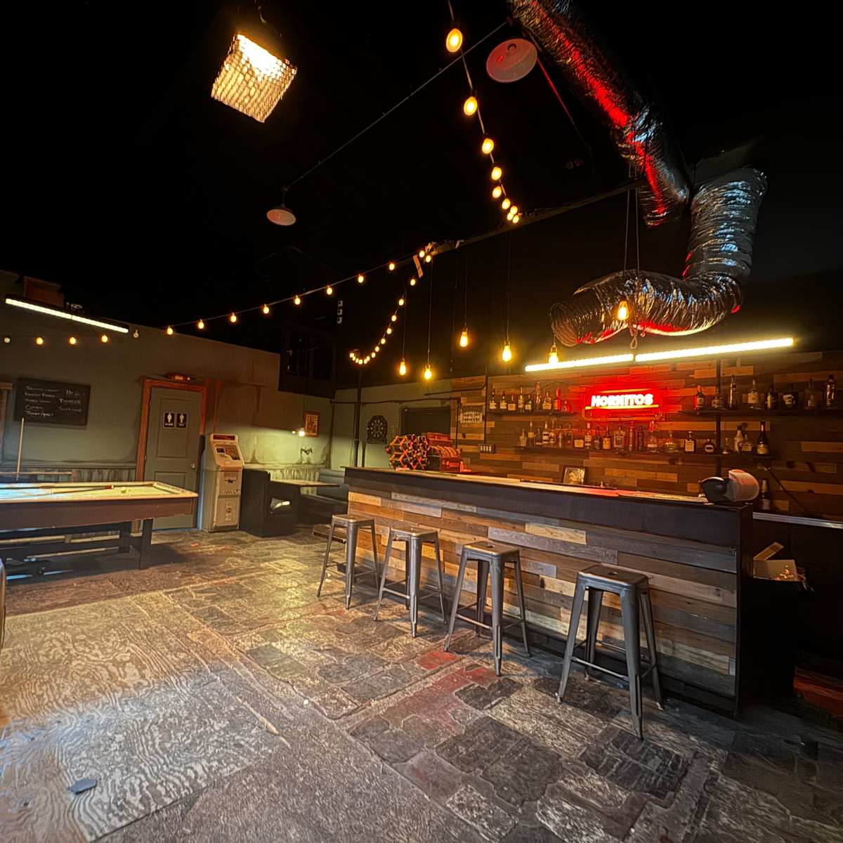 An interior of a bar or game room with a pool table, a wooden bar counter, and string lights hanging from the ceiling.