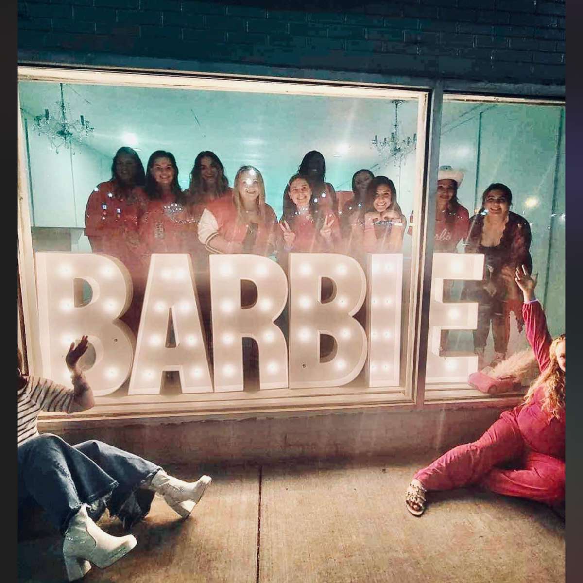 A group of women poses together in a storefront window beside large illuminated letters spelling "BARBIE."