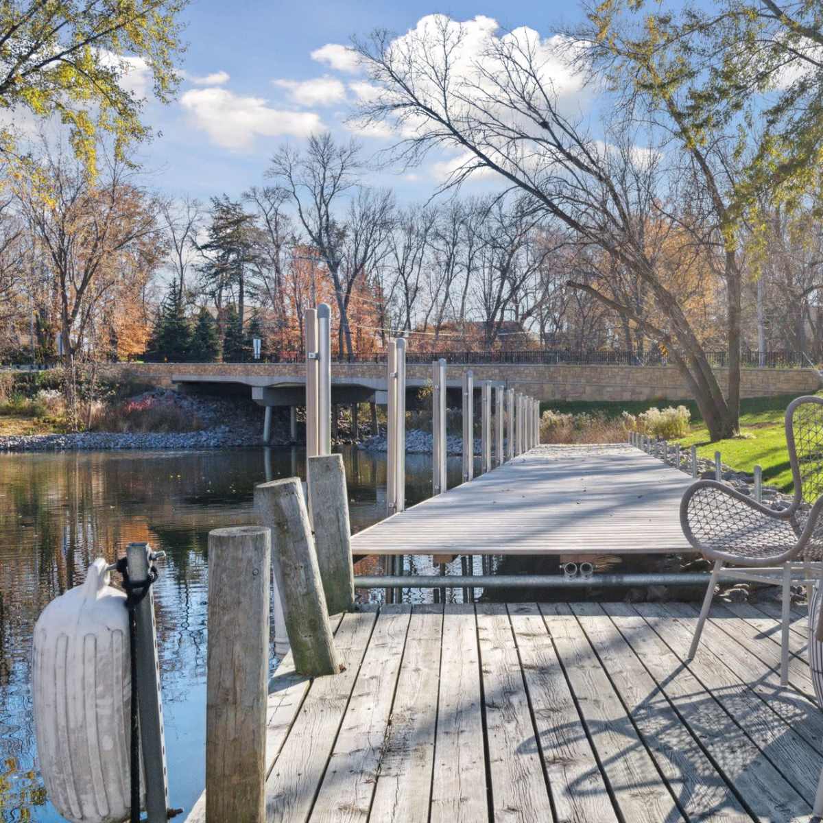 A wooden dock extends over a calm waterway, lined with chairs and surrounded by trees and a bridge in the background.