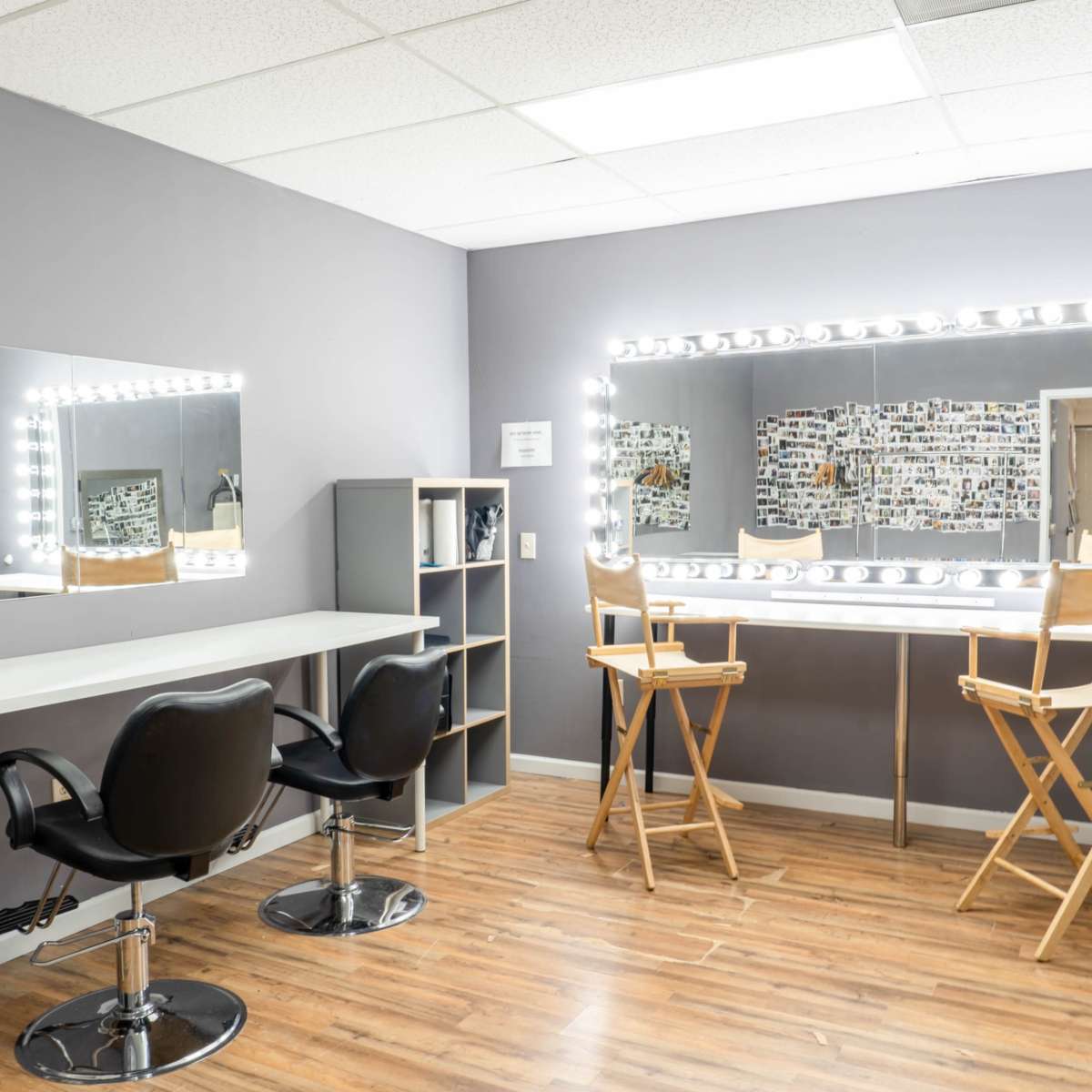 A mirror-lined makeup station with two chairs and a shelving unit in a room with gray walls and wooden flooring.
