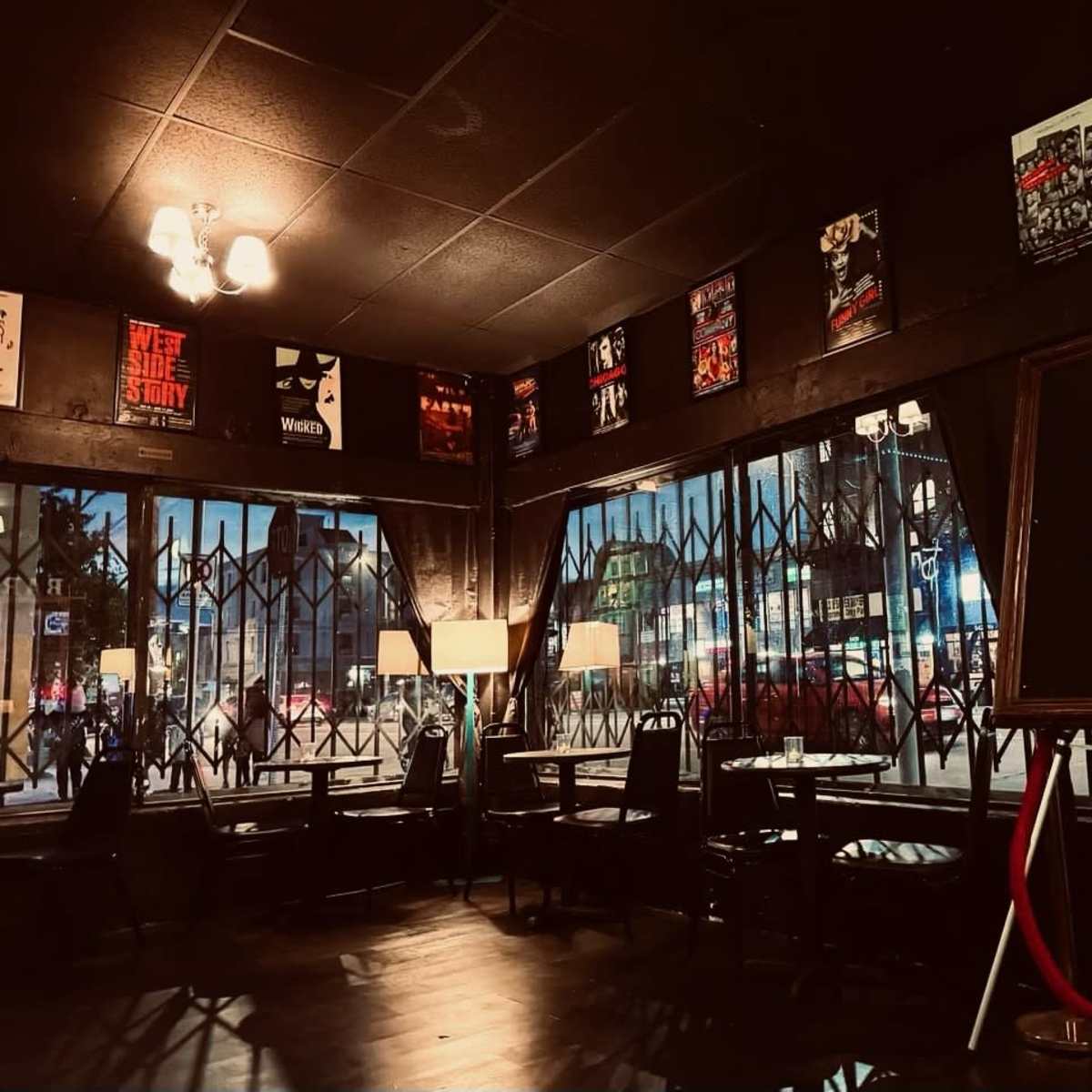 The interior of a dimly lit restaurant features black walls adorned with posters, and large windows secured with metal grilles showing street scenes outside.