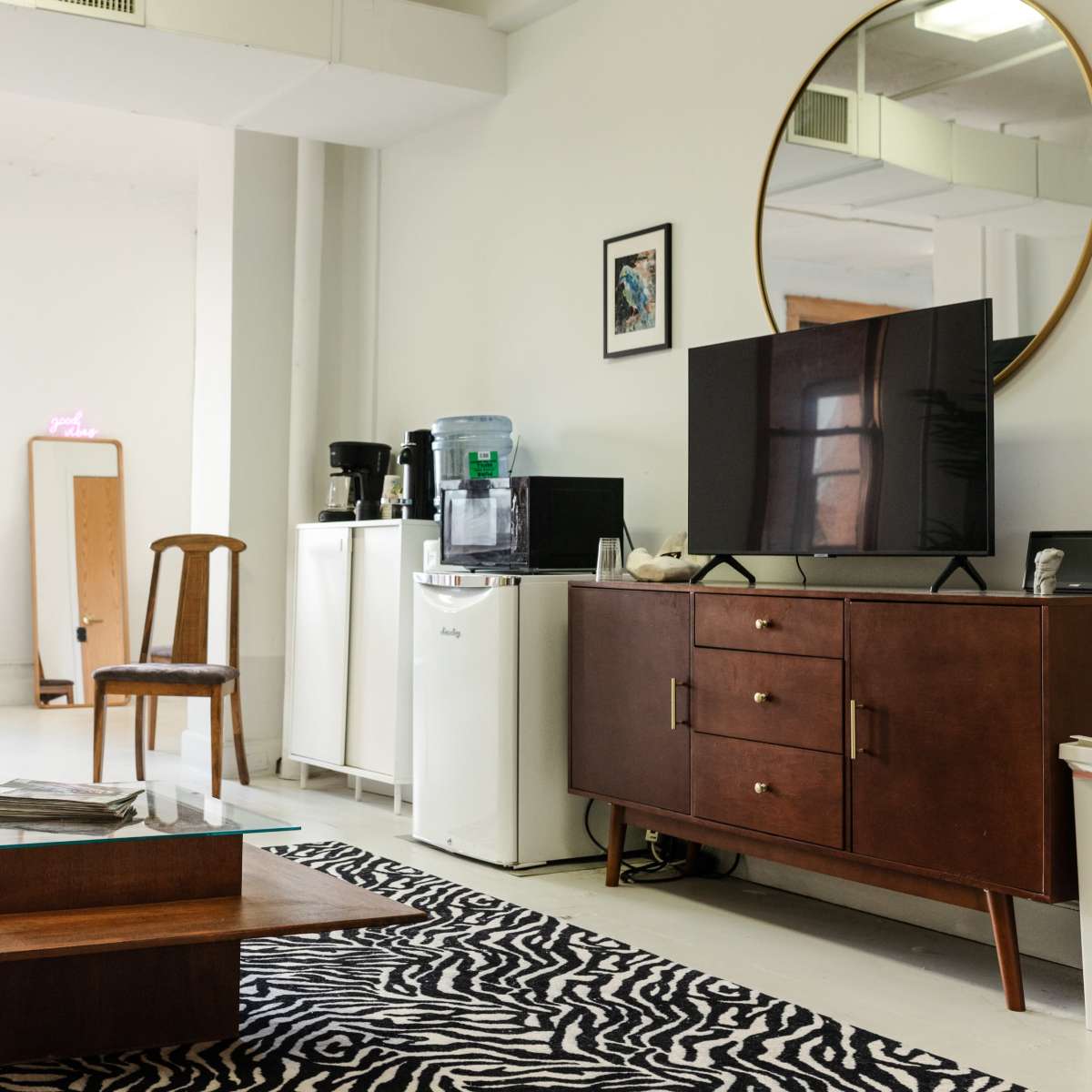 A modern, furnished living room with a large mirror, a television on a wooden cabinet, a coffee maker, and a table on a zebra-striped rug.