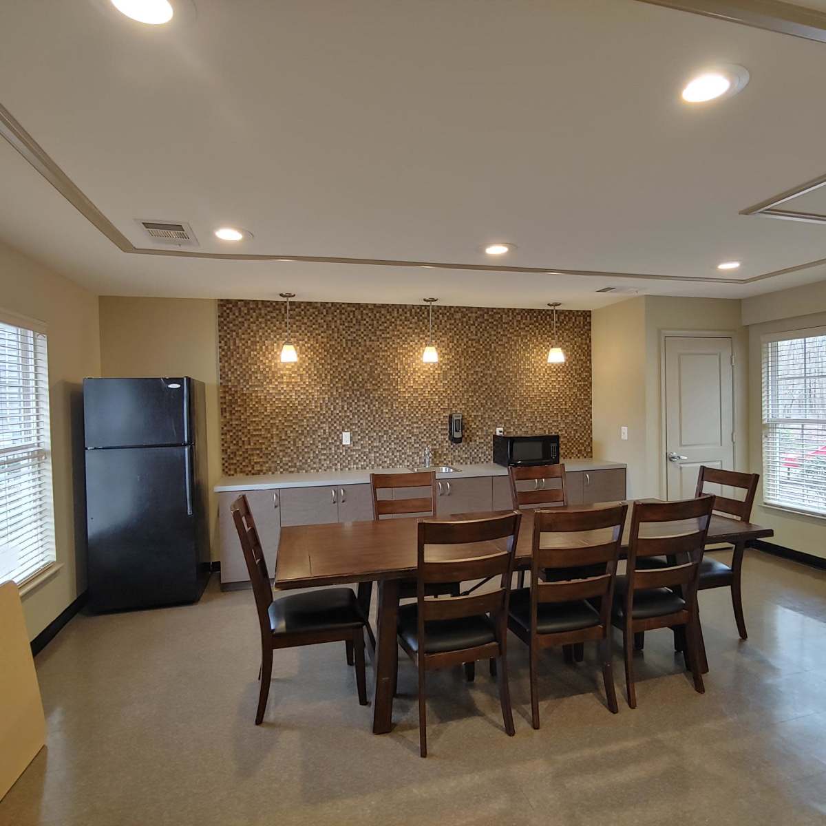 A modern kitchen dining area features a large wooden table with eight chairs, a refrigerator, a microwave, and decorative wall paneling.