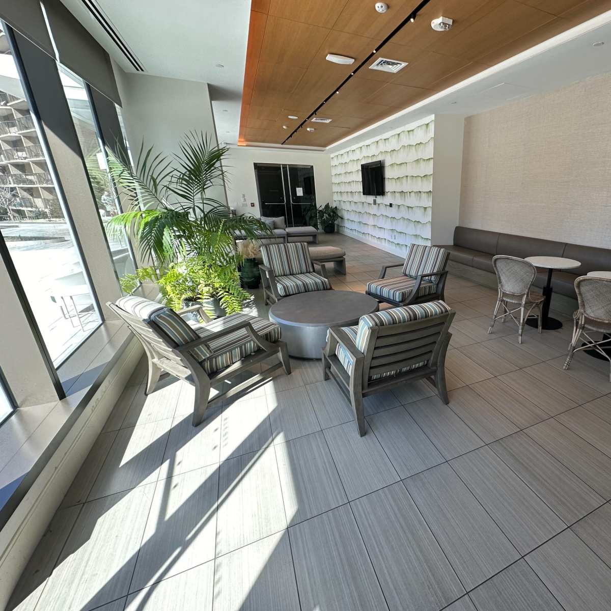 A modern lounge area with several seating arrangements, including striped chairs and small round tables, surrounded by greenery and large windows.