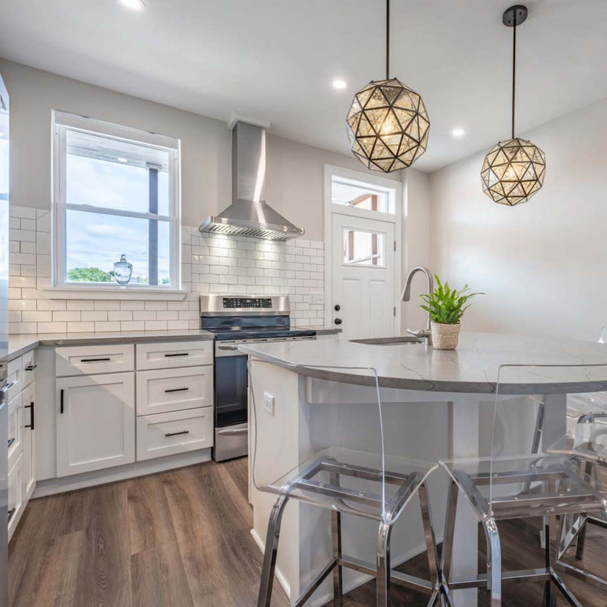 A modern kitchen features a round island with transparent stools, pendant lighting, stainless steel appliances, and a window above the sink.