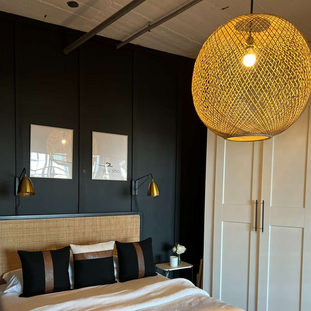 A modern bedroom featuring a bed with a white quilted coverlet, black pillows, a woven pendant light, and a black accent wall with two framed artworks.