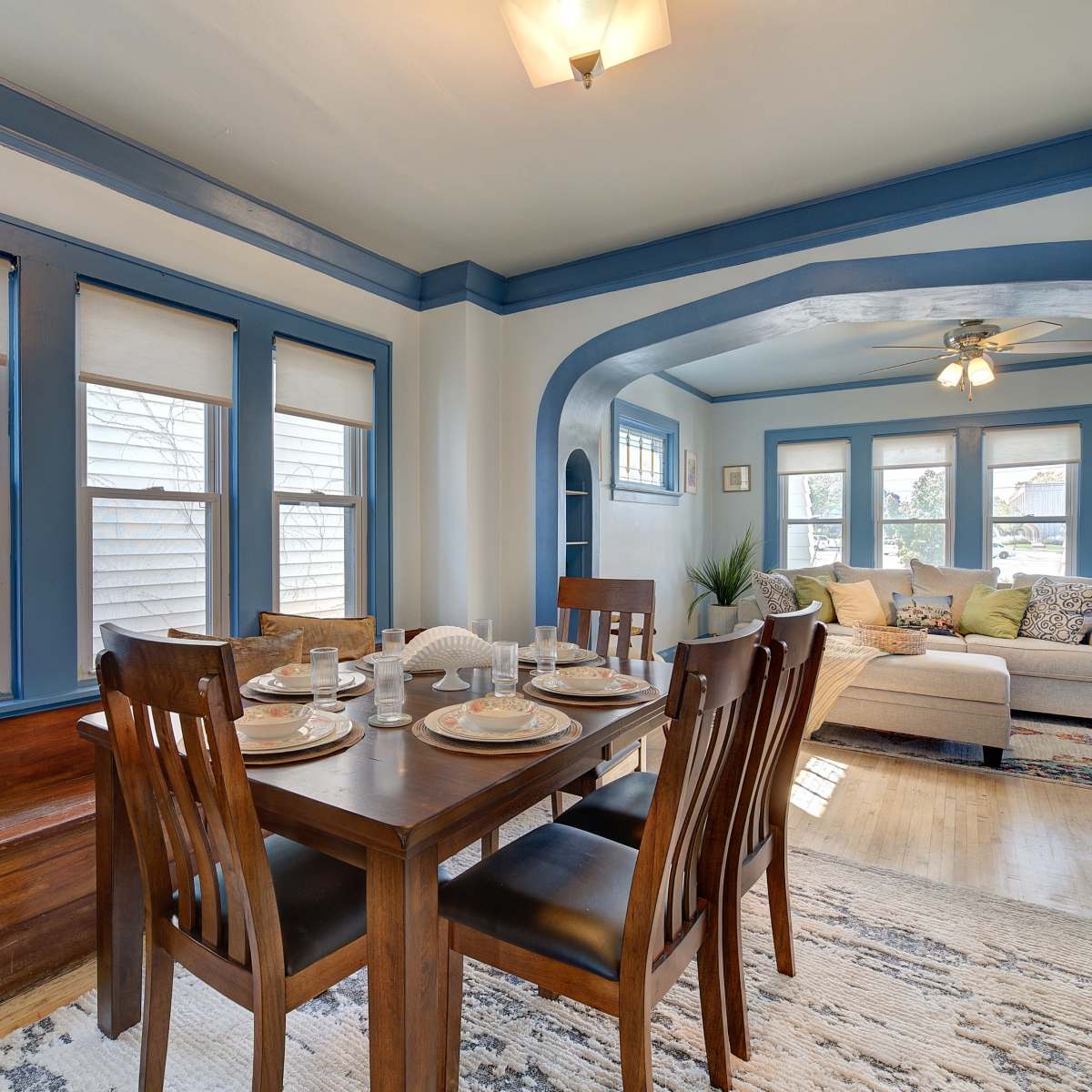 A dining area with a wooden table set for four, adjacent to a living room featuring a light-colored sofa and several windows.