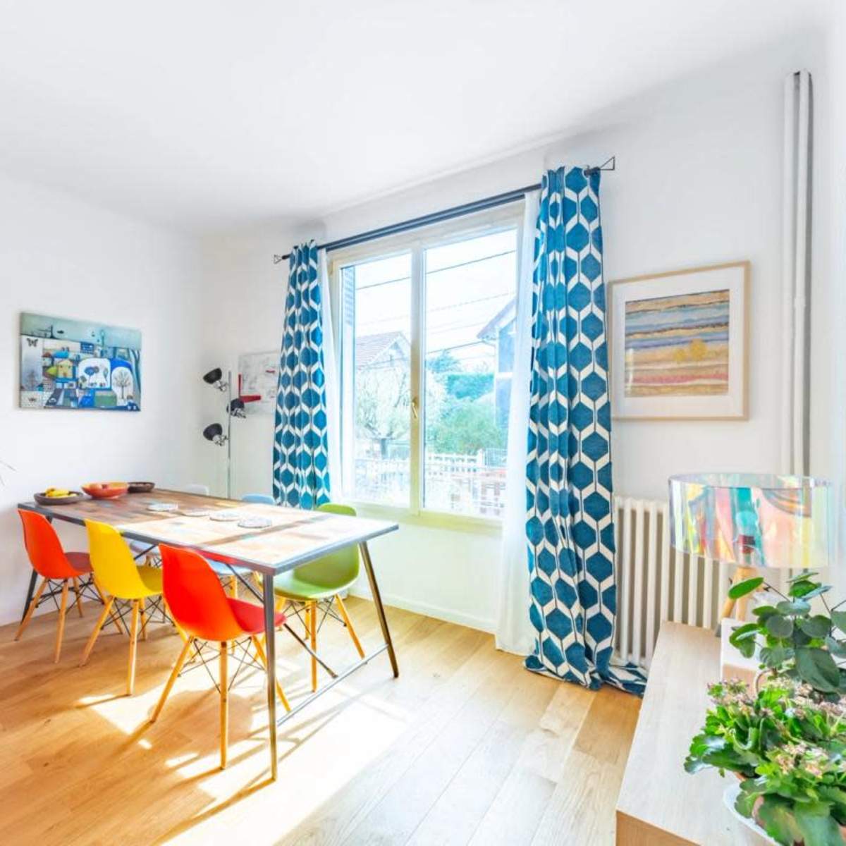 A dining area features a table surrounded by colorful chairs, with a large window letting in natural light and a potted plant in the corner.