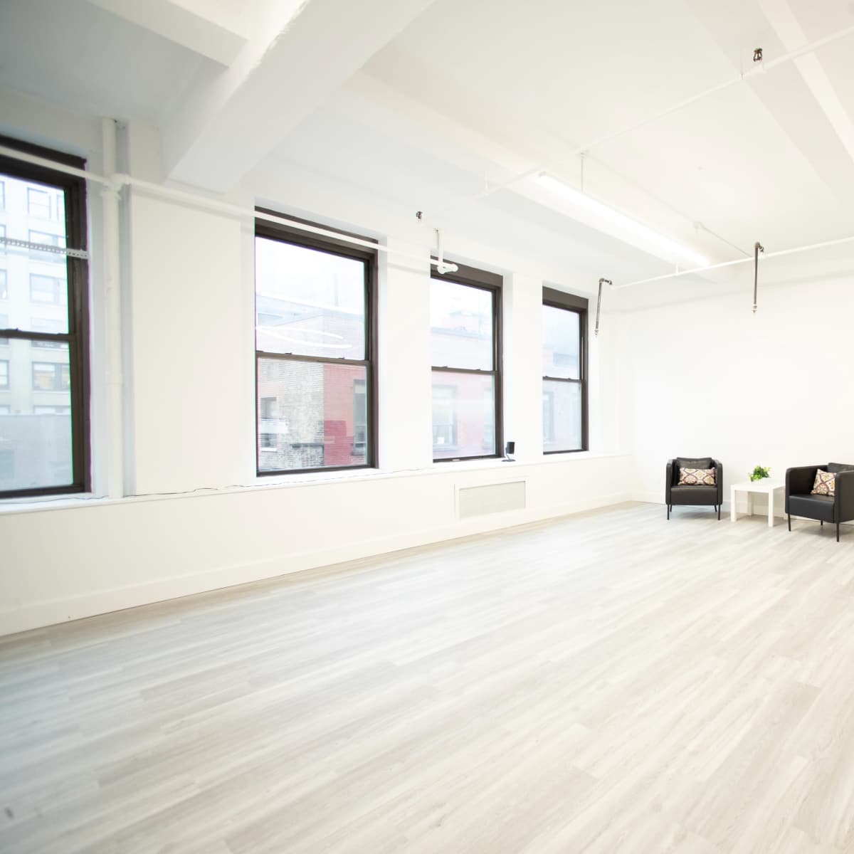 A spacious, minimally furnished room with two black chairs, a small table, and a desk against a white wall, illuminated by natural light from large windows.