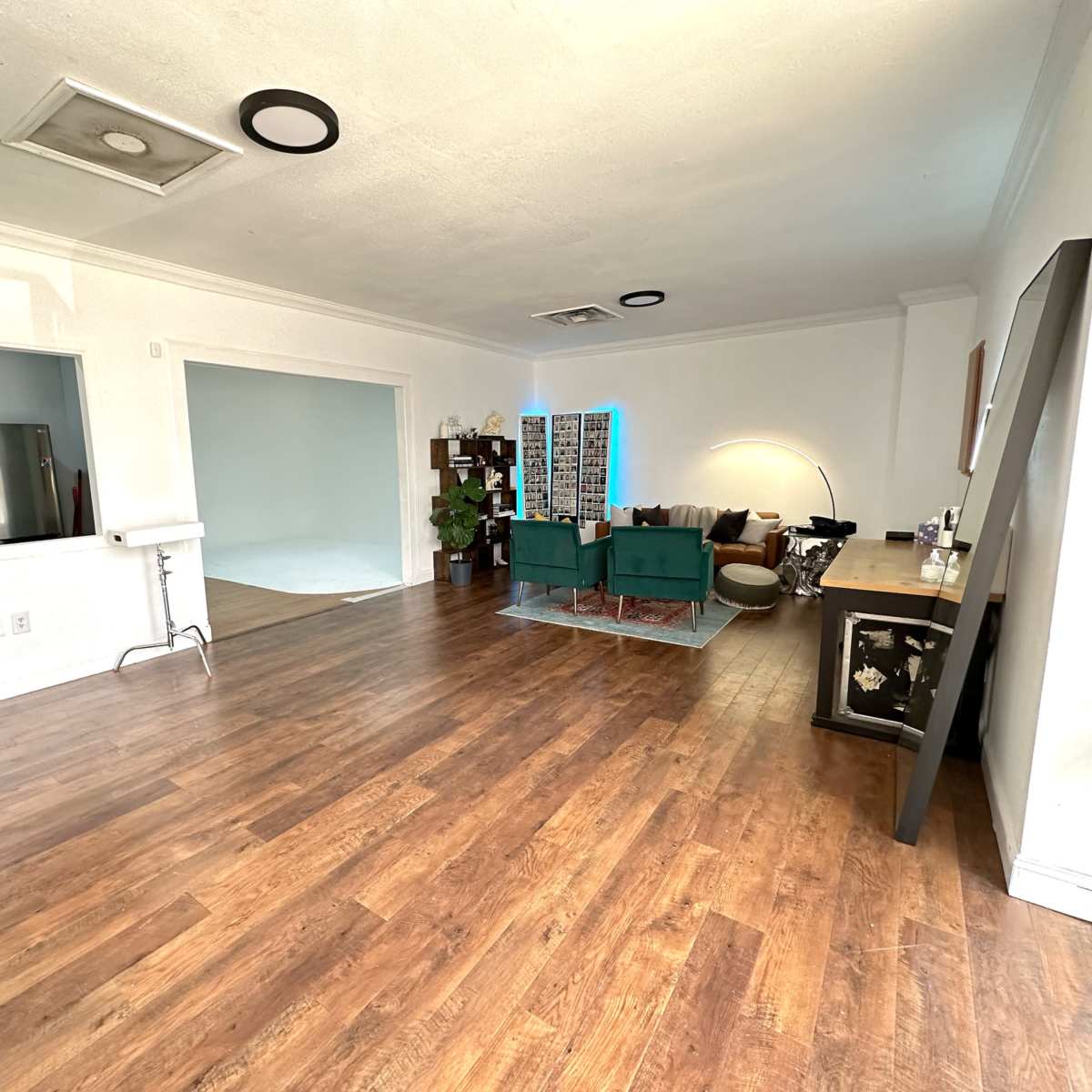 A spacious, well-lit room with wooden flooring, featuring a kitchen area on the left and a seating area with green chairs and a light-colored wall in the background.