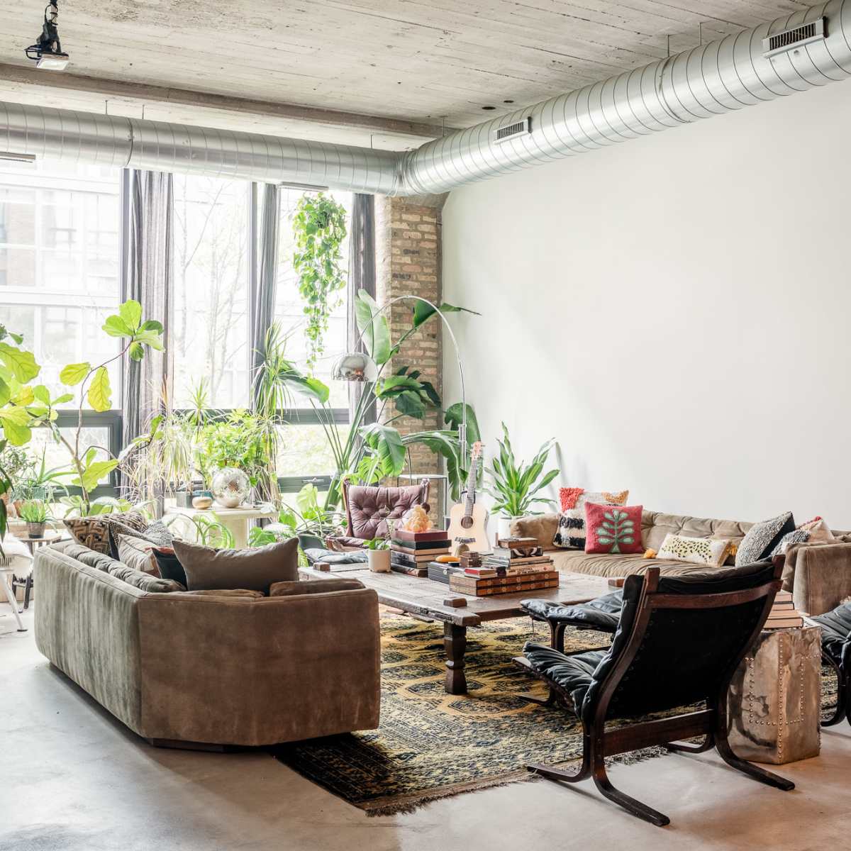 A spacious living room features a large sectional sofa, a coffee table surrounded by books and plants, and ample natural light from large windows.