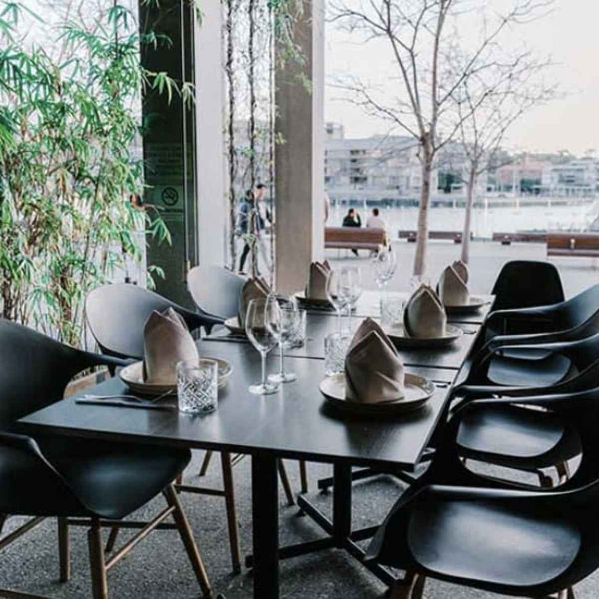 A dining table is set with glassware and folded napkins in a modern restaurant overlooking a body of water.