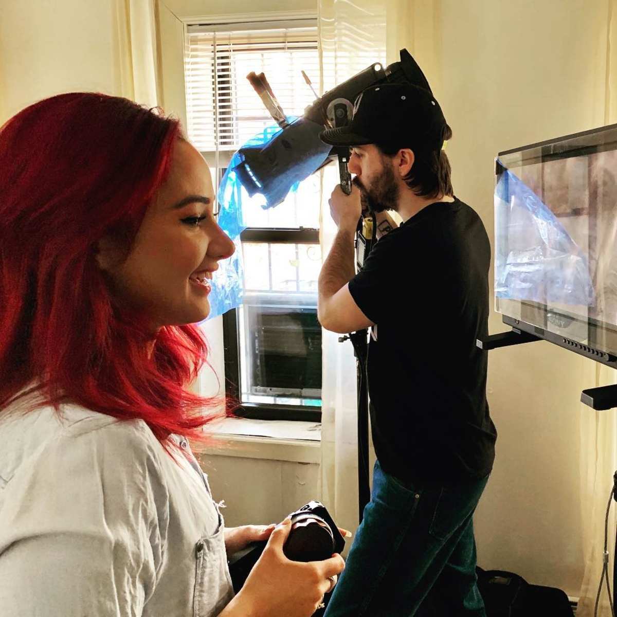 A woman with red hair smiles while a man behind her operates a camera near a monitor in a well-lit room.