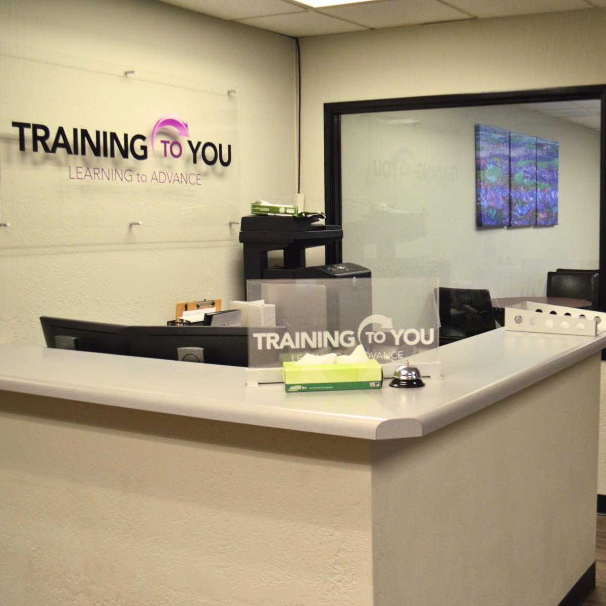 A reception area with a gray counter and a glass wall leading to a conference room, featuring a logo that reads "TRAINING TO YOU."