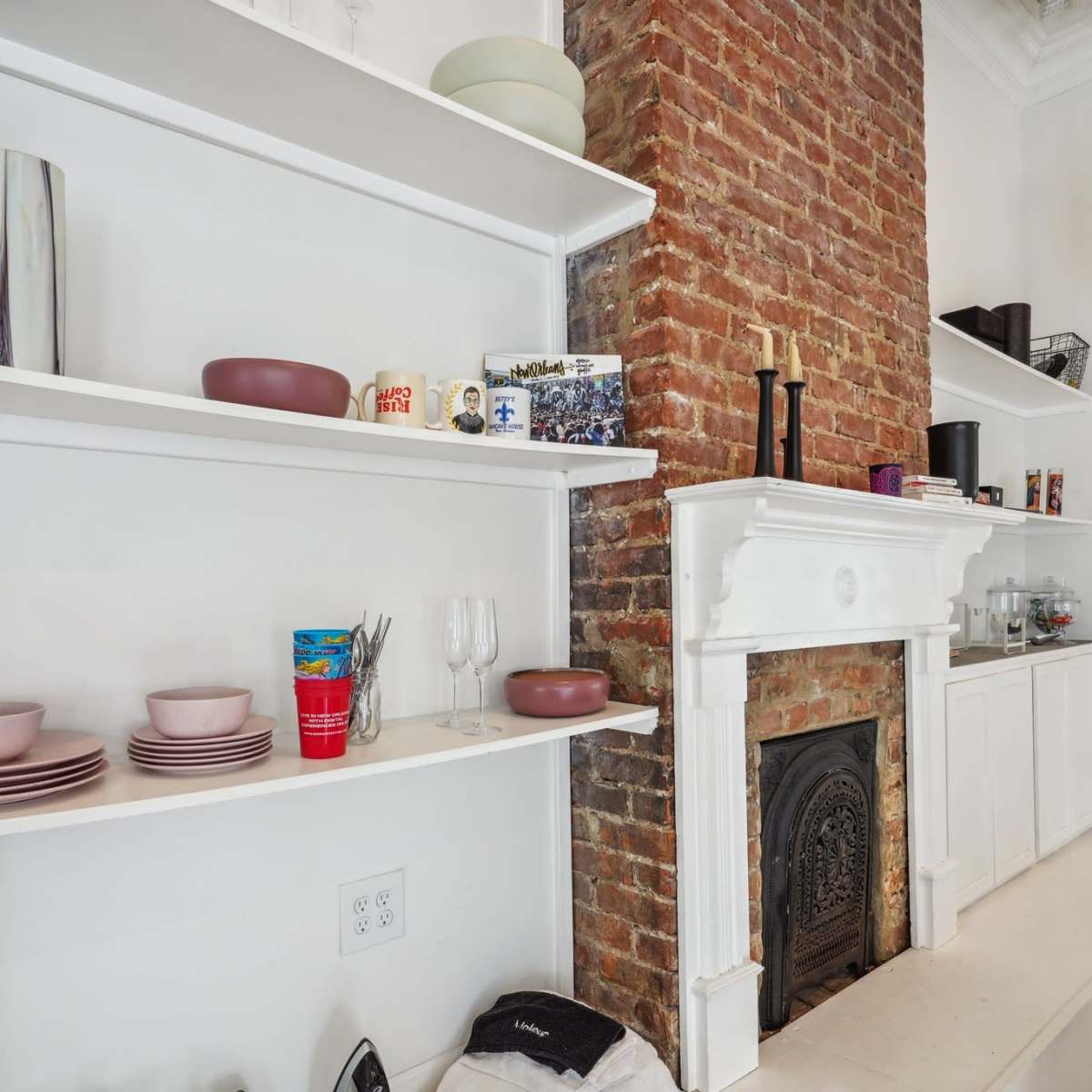A white shelf displaying various dishes and glassware next to a brick fireplace with a white mantel in a bright room.
