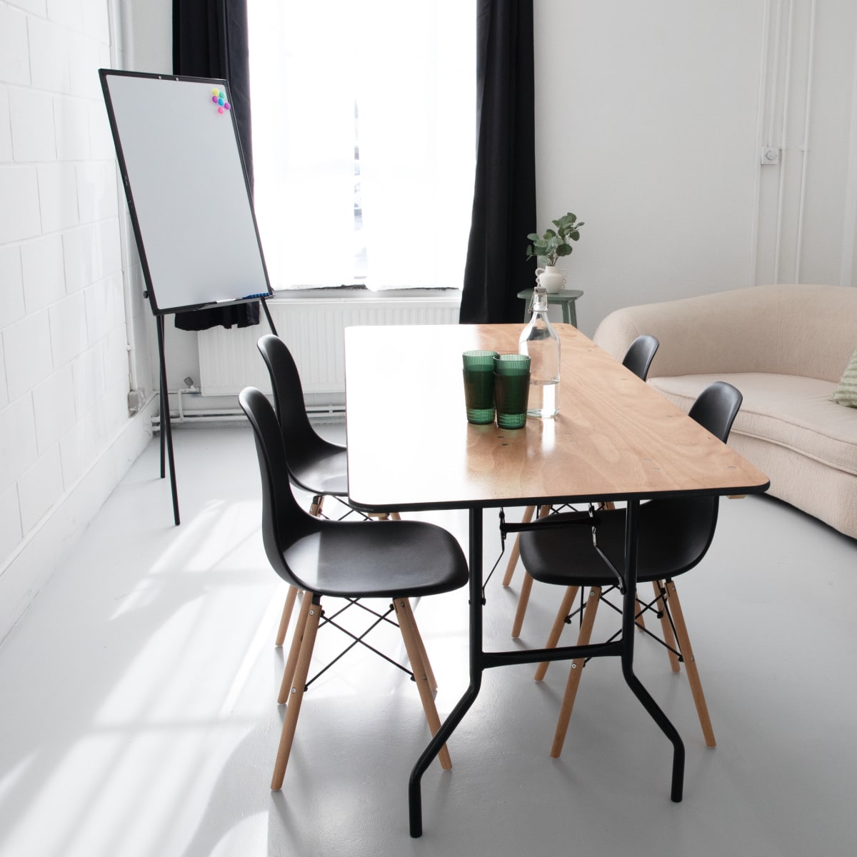 A minimalist meeting room features a wooden table surrounded by black chairs, with a whiteboard and a couch in the background.