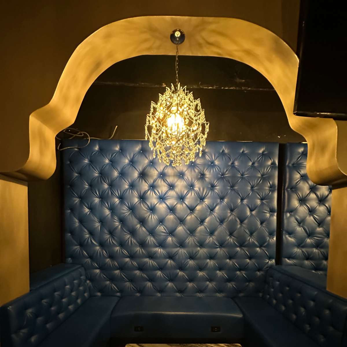 A dimly lit lounge area with a blue tufted seating arrangement, a marble table, and a crystal chandelier hanging from the ceiling.
