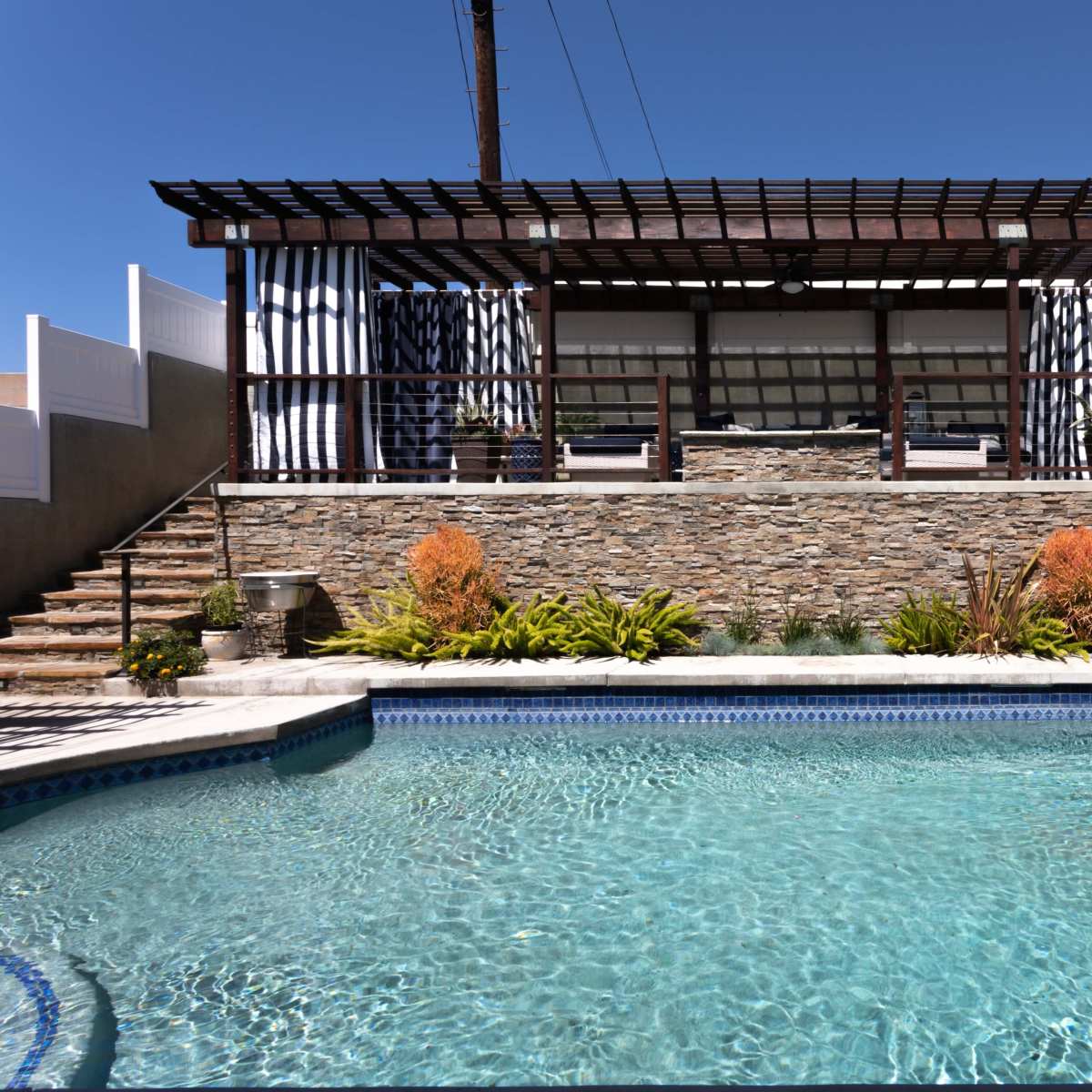 A swimming pool with a stone terrace and a pergola in the background, surrounded by landscaped plants and stairs.