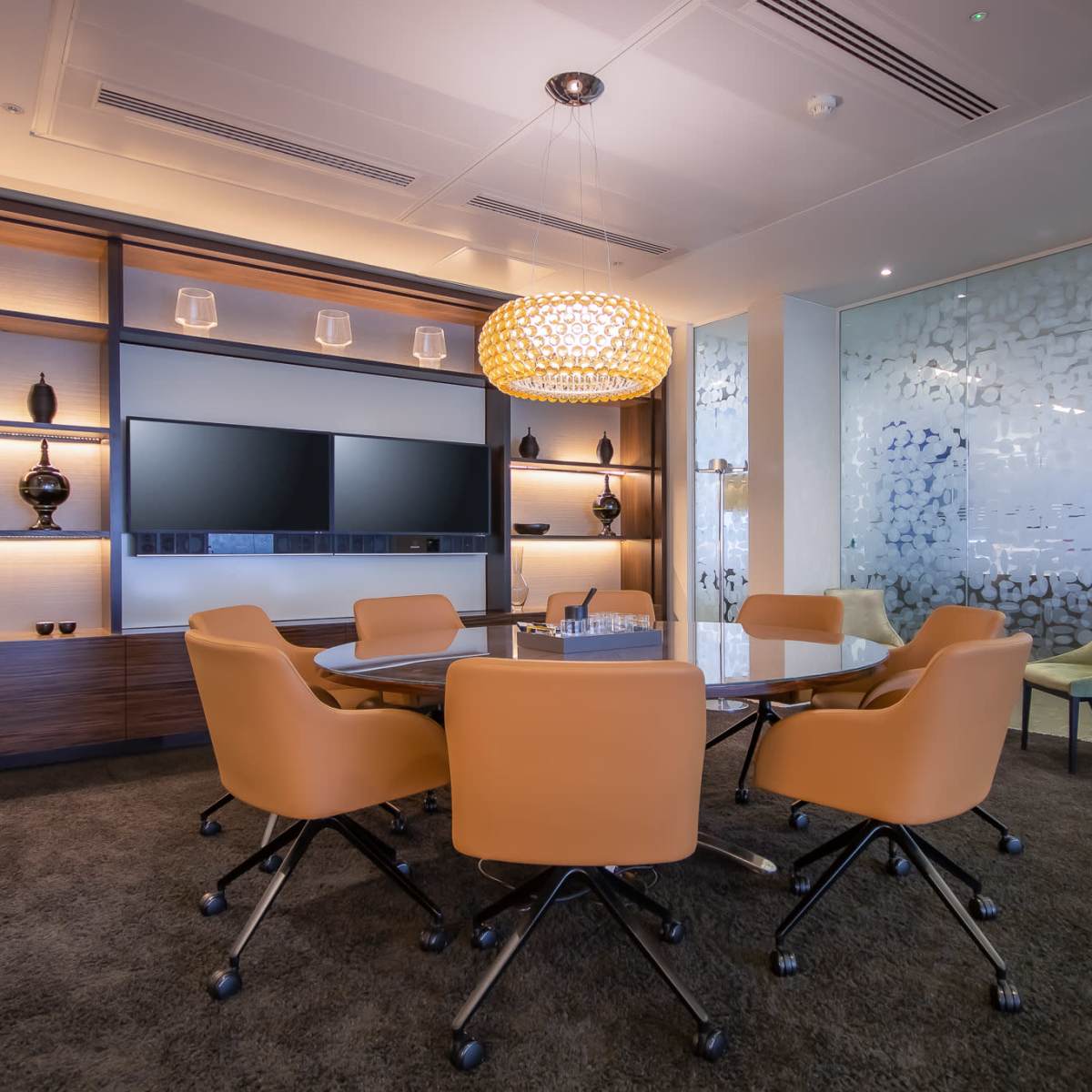 A modern conference room features a round table surrounded by orange chairs, with a display screen and decorative shelving in the background.