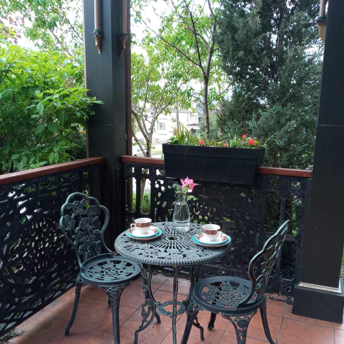 A small metal table with two chairs is set on a balcony surrounded by greenery, featuring two cups and a flower in the center.