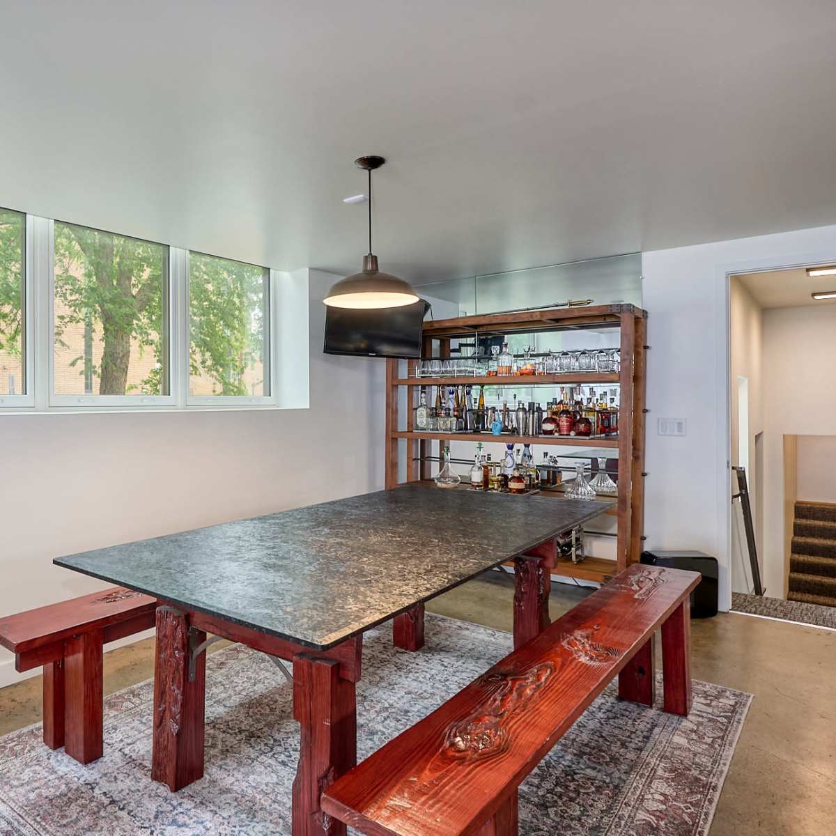 A modern dining area featuring a large black table with red wooden benches, a shelf with bottles in the background, and windows allowing natural light to enter.