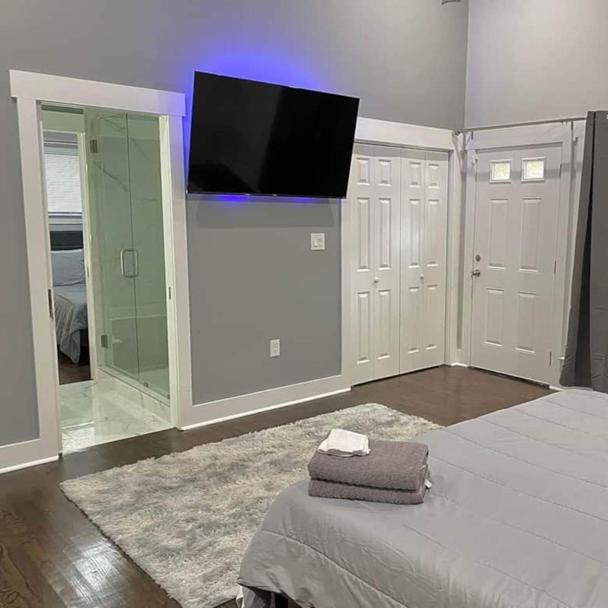 A modern bedroom with a large bed covered in gray bedding, a flat-screen TV mounted on the wall, a bathroom visible through a glass door, and gray curtains covering a window.