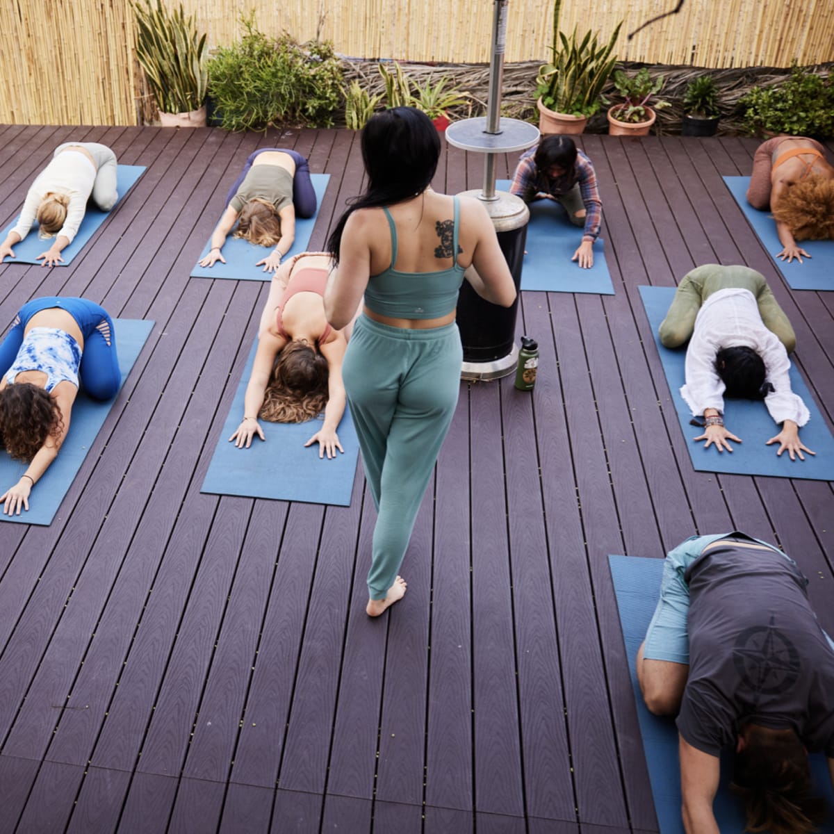 A group of people practice yoga on mats in an outdoor studio with bamboo fencing and potted plants.