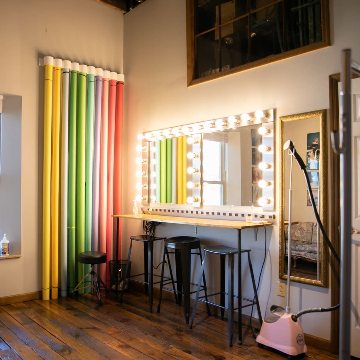 A brightly lit makeup area with a large mirror surrounded by bulbs, a wooden floor, and colorful backdrop rolls.