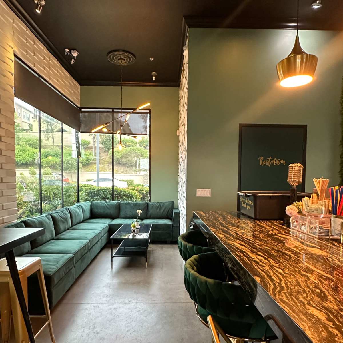 A modern cafe interior featuring a green velvet seating area, a marble countertop bar, and large windows overlooking an outdoor space.