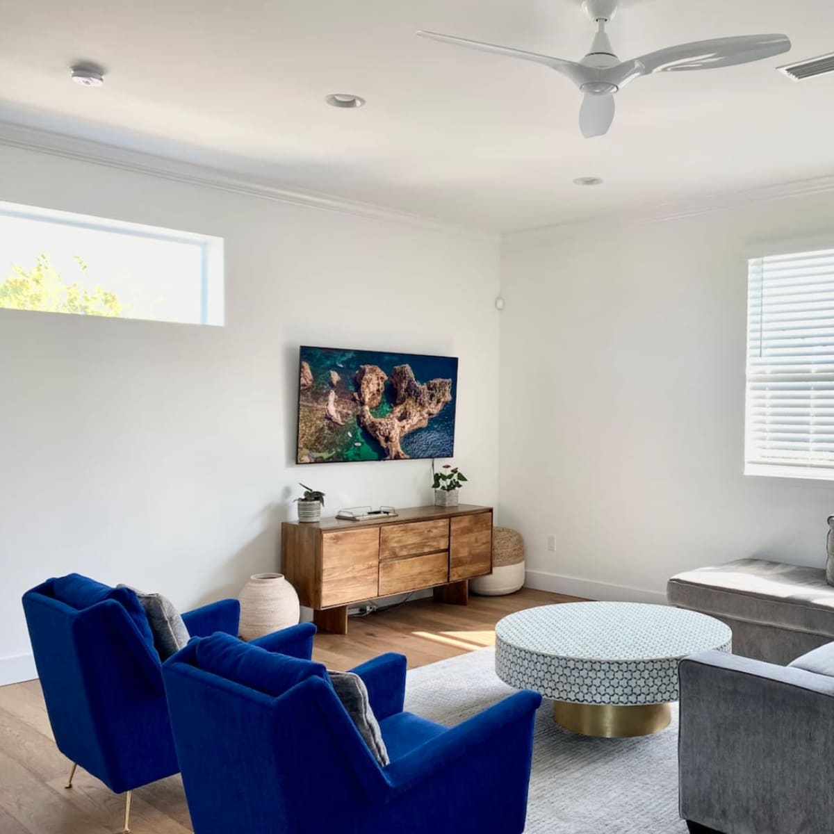 A modern living room with a sectional sofa, two blue chairs, a round coffee table, and a wall-mounted TV above a wooden console.
