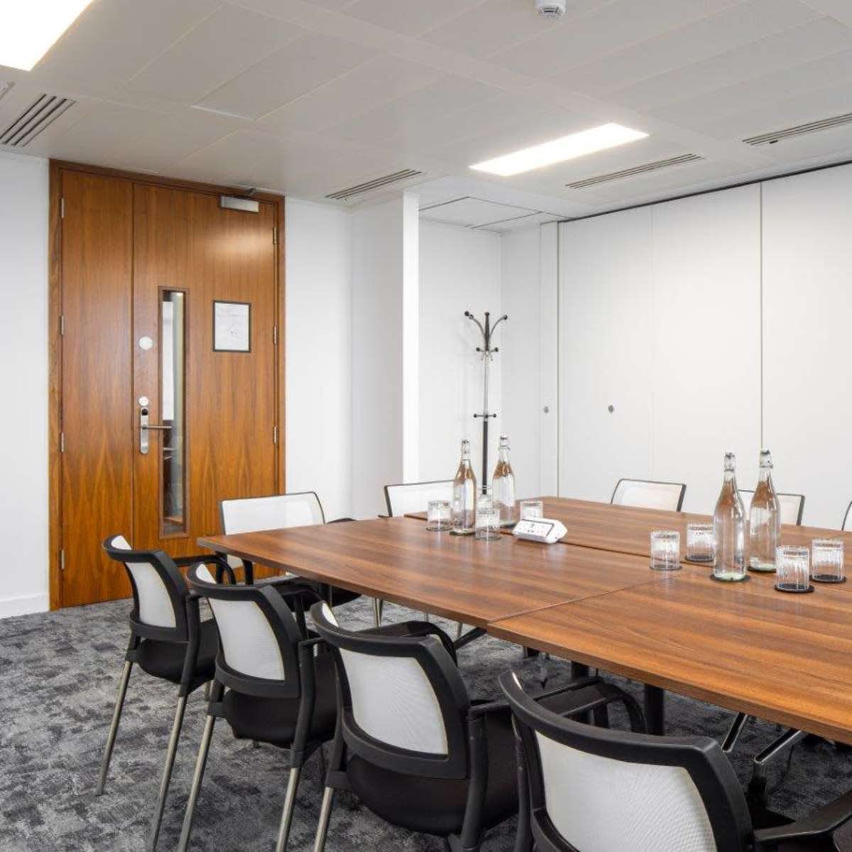 A large meeting room features a long wooden table surrounded by chairs, with water bottles and glasses placed on the table.