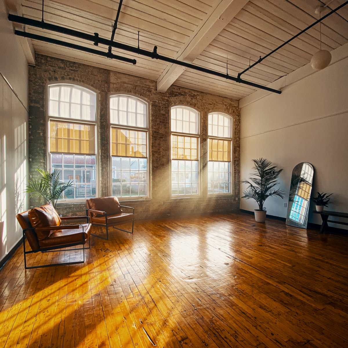 A spacious room with large windows, wooden flooring, and two leather chairs, illuminated by sunlight streaming through the windows.