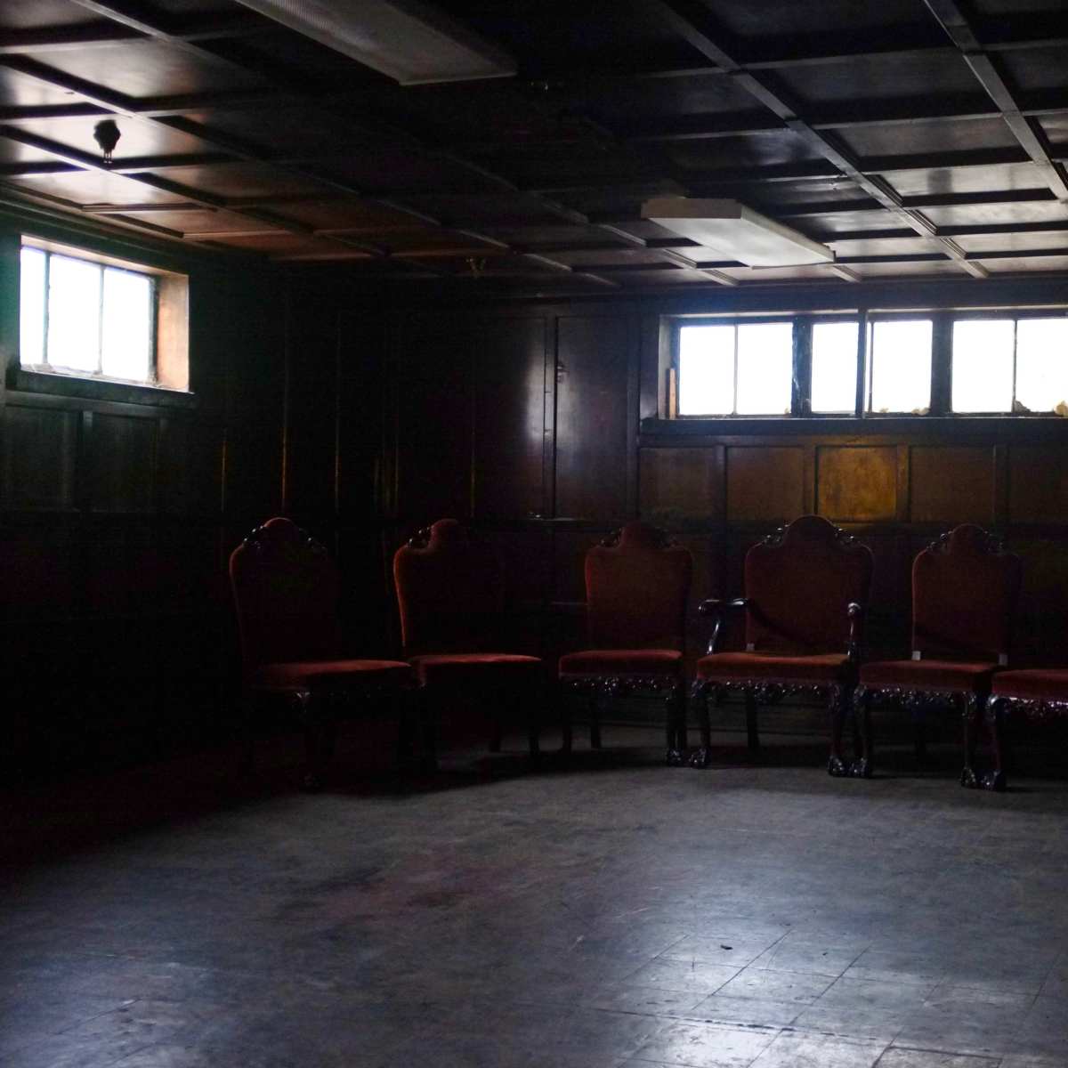 A dimly lit room features a row of five ornate chairs against a dark wooden wall, with two small windows allowing faint light to enter.