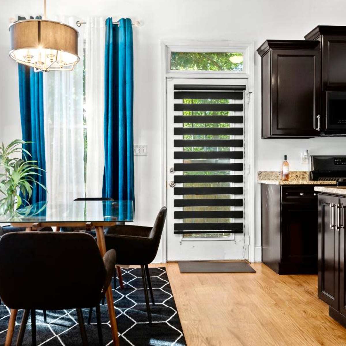 A kitchen with dark wood cabinets, a glass-top dining table, four black chairs, and large windows covered with blue curtains.