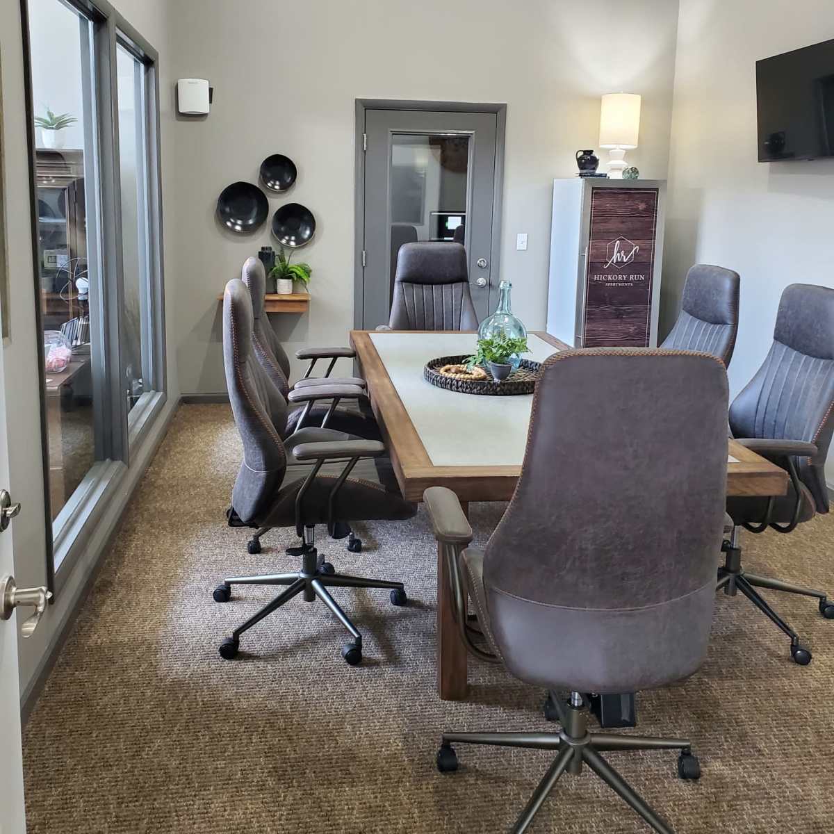 A conference room features a large rectangular table surrounded by six ergonomic chairs, with a side door and windows allowing natural light.