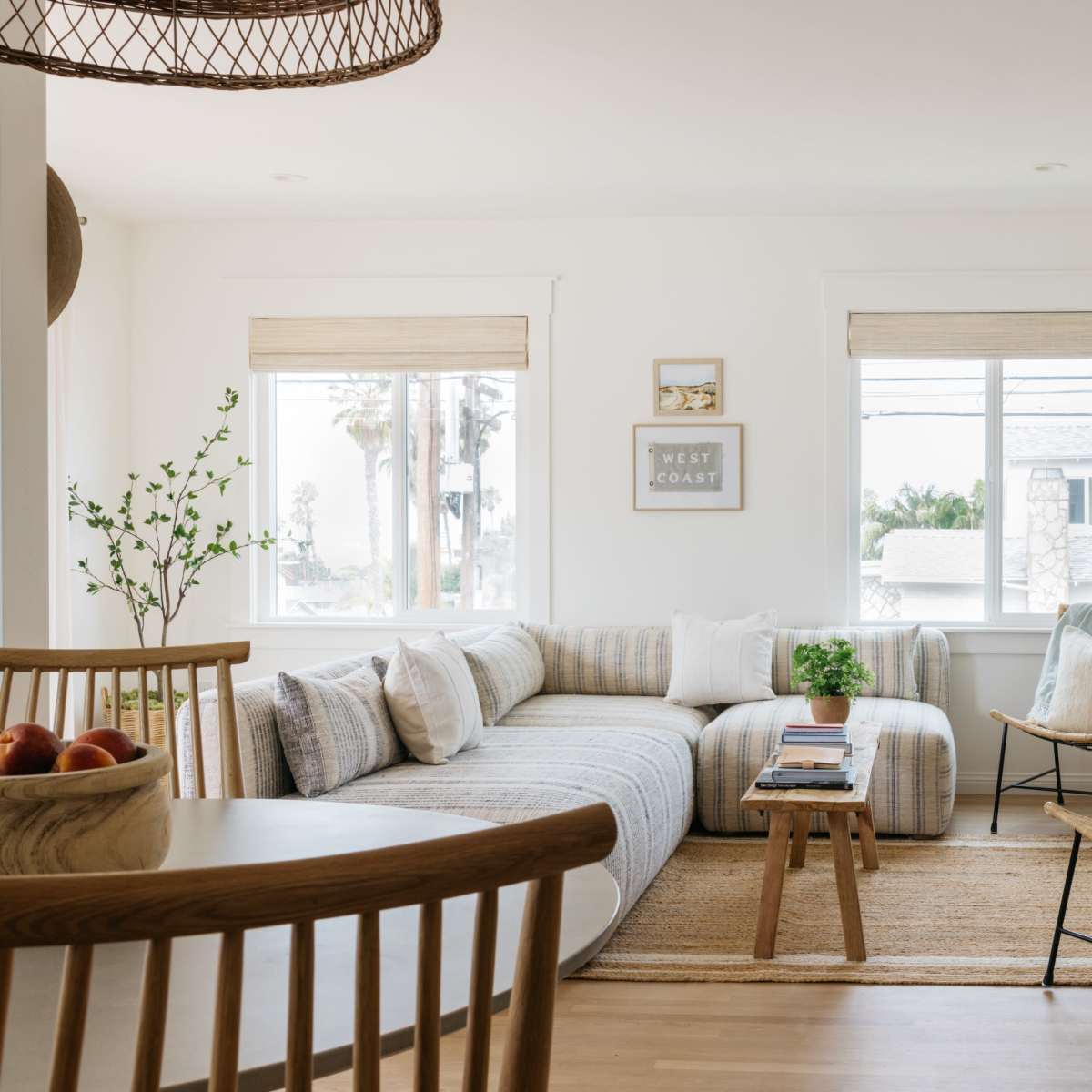 A bright living room features a sectional sofa with striped fabric, a wooden coffee table, and a mix of chairs, all arranged near large windows with natural light streaming in.