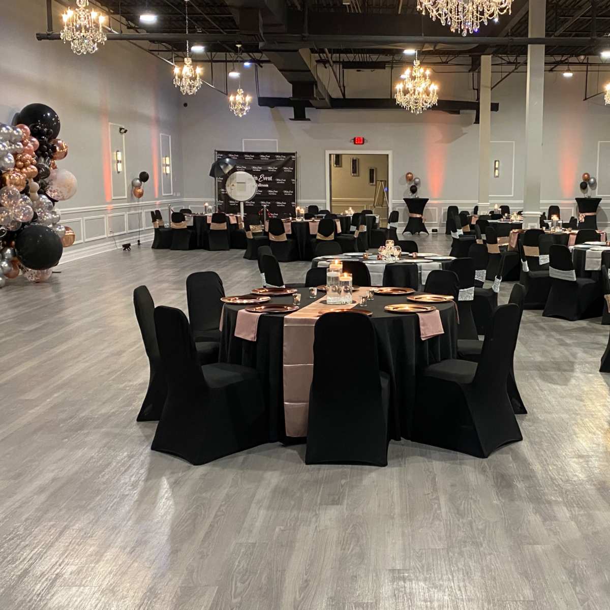A spacious banquet hall with elegantly arranged tables covered in black and rose gold linens, surrounded by decorative chandeliers.