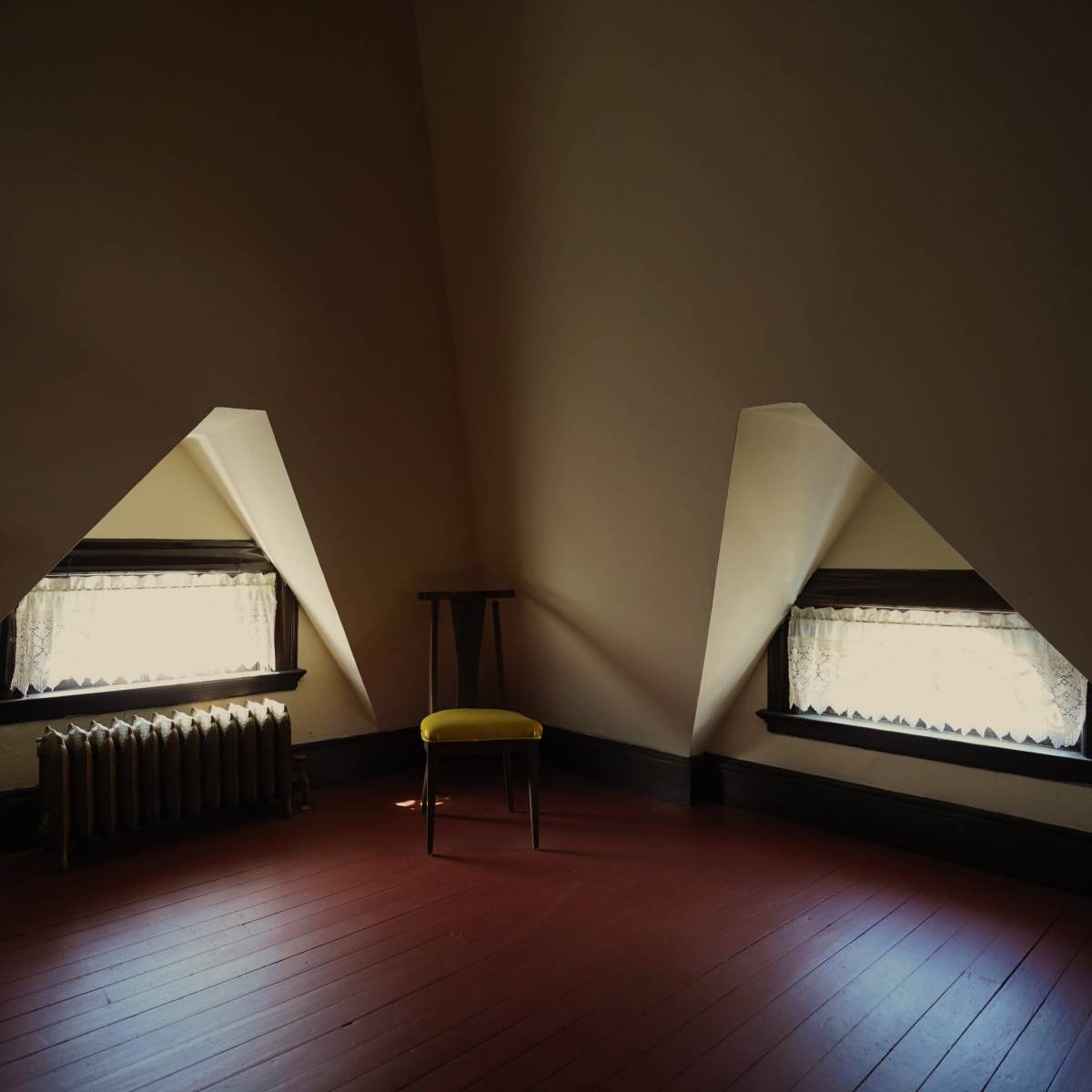 A small, sparsely furnished attic room features two triangular windows, a wooden chair, and a radiator along the wall.
