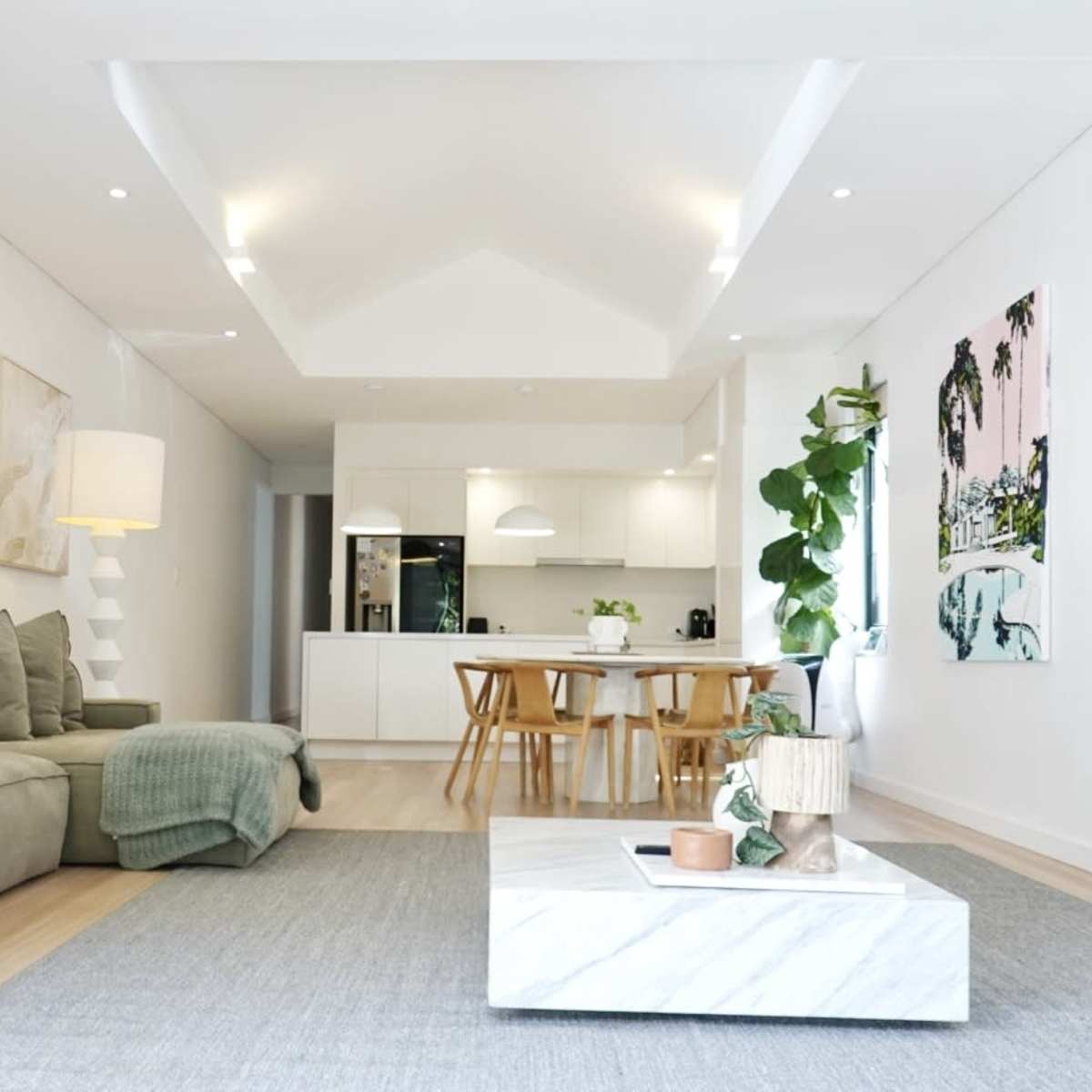 A modern living room with a sectional sofa, a marble coffee table, and a dining area featuring wooden chairs and a kitchen in the background.