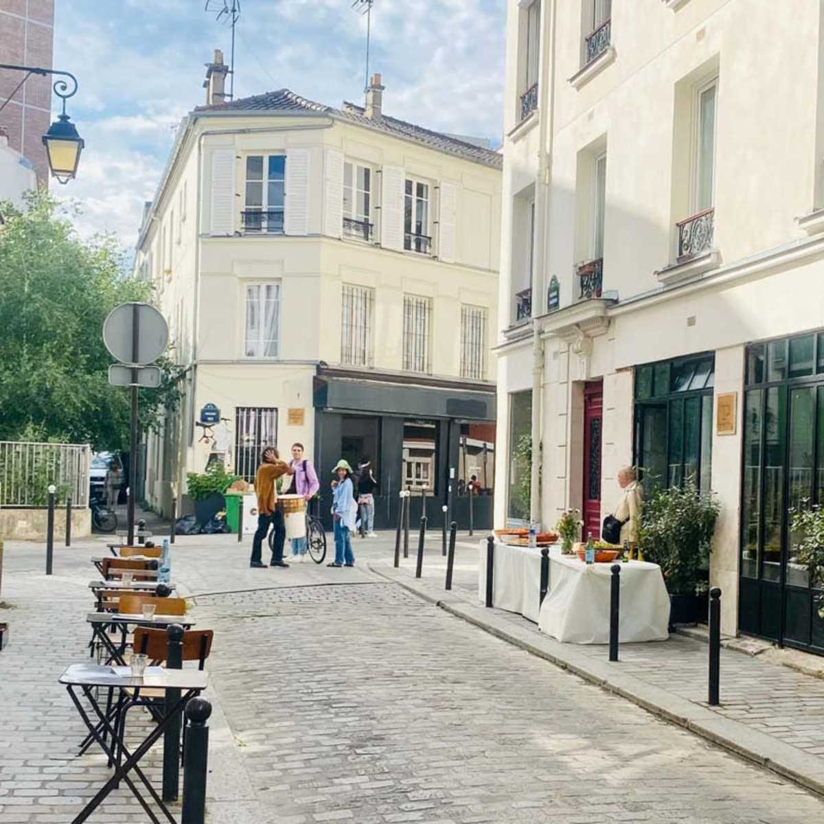 A cobblestone street is lined with small tables and chairs outside a café, with people gathered and a decorative table set up nearby.