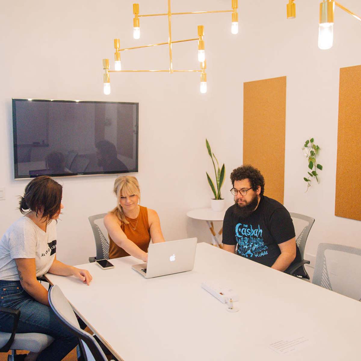 Three people sit at a modern conference table working on a laptop in a well-lit meeting room.